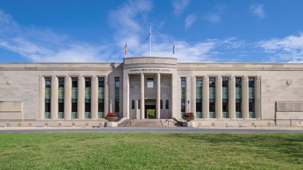 Art Deco stone building facade with signage reading "National Film and Sound Archive", with bright green grass foreground and bright blue sky background