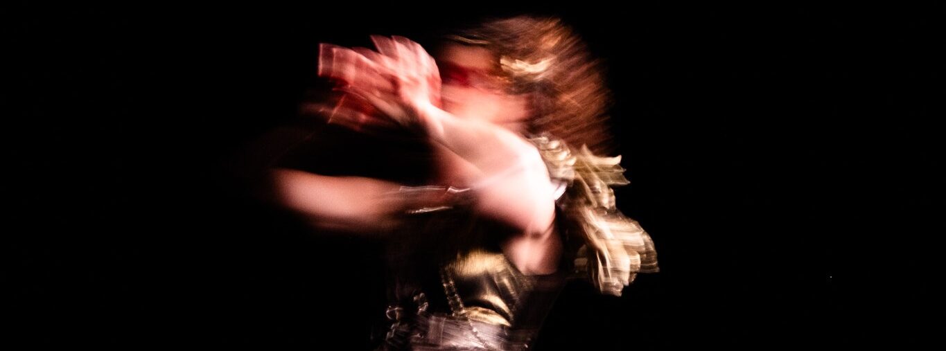 A blurred image of a woman in a gold costume signing, in profile against a black background