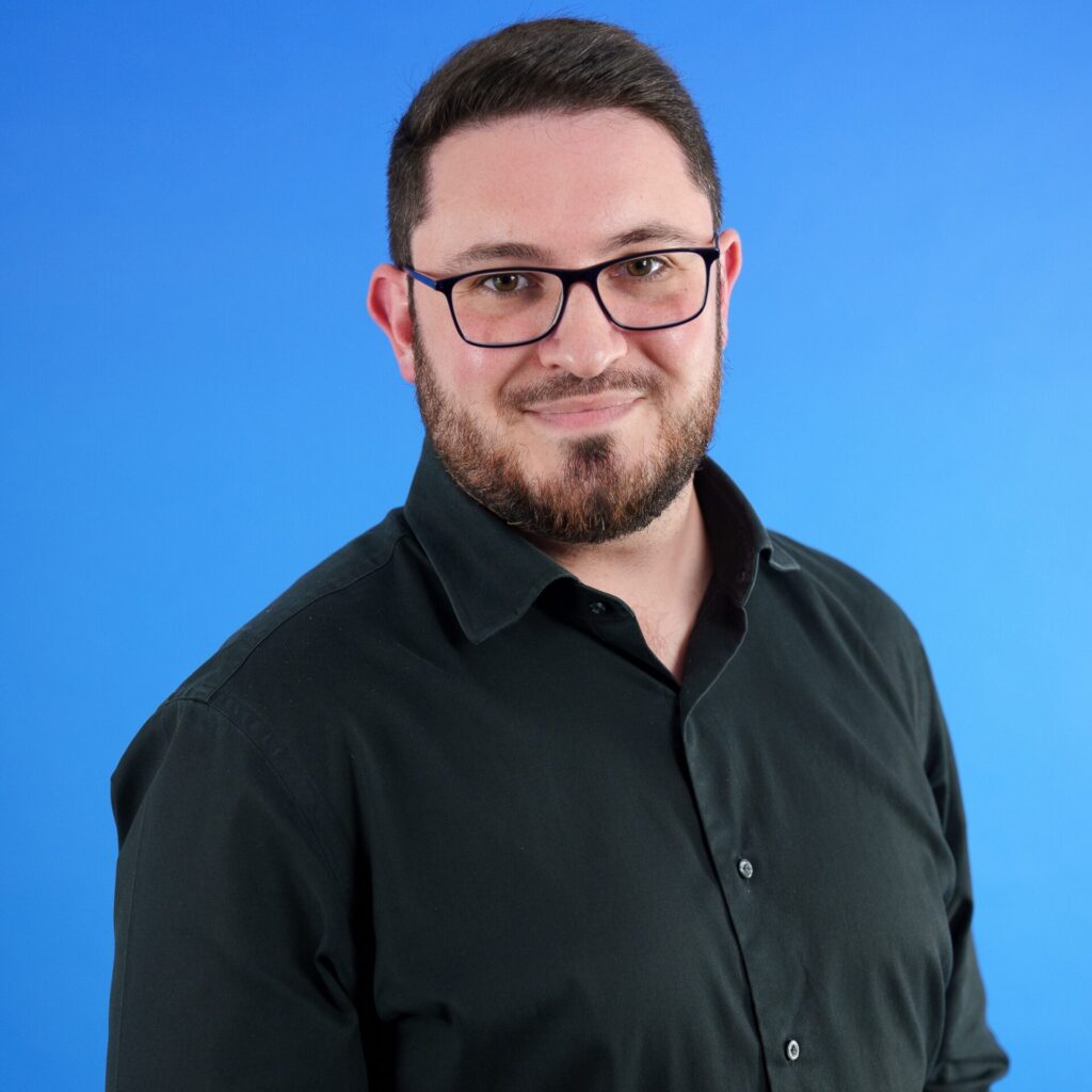 Bearded man with glasses smiles against a blue background