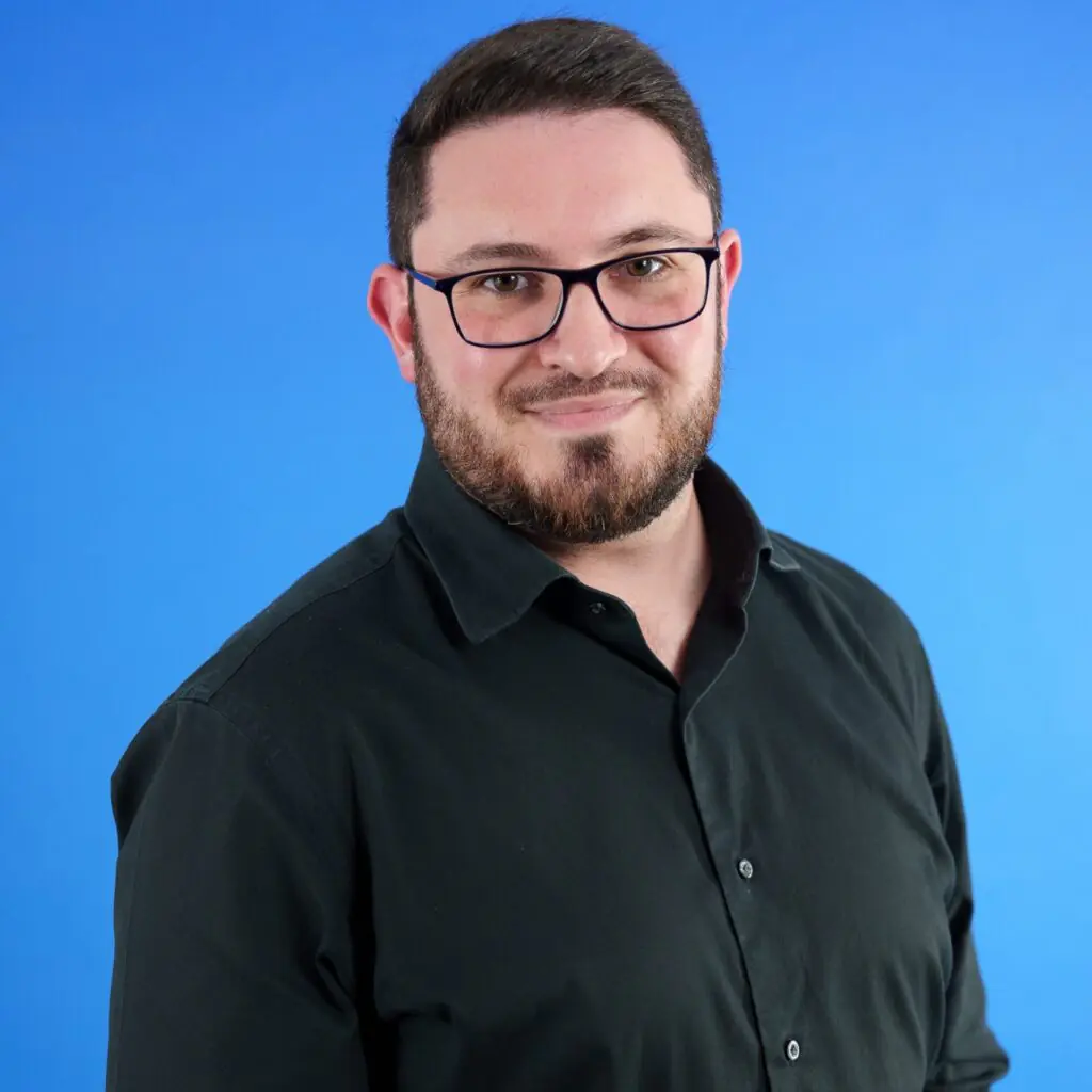 Bearded man with glasses smiles against a blue background