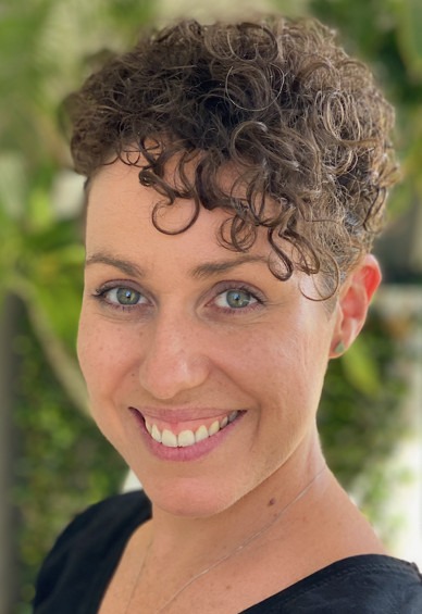 Woman with curly brown hair smiles against a leafy background