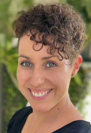 Woman with curly brown hair smiles against a leafy background