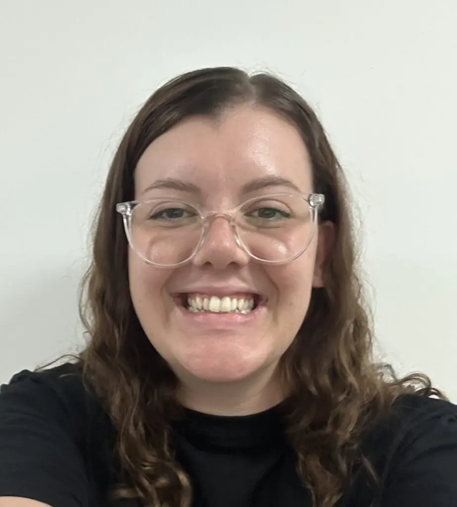Woman with curly hair and glasses smiles against a grey background