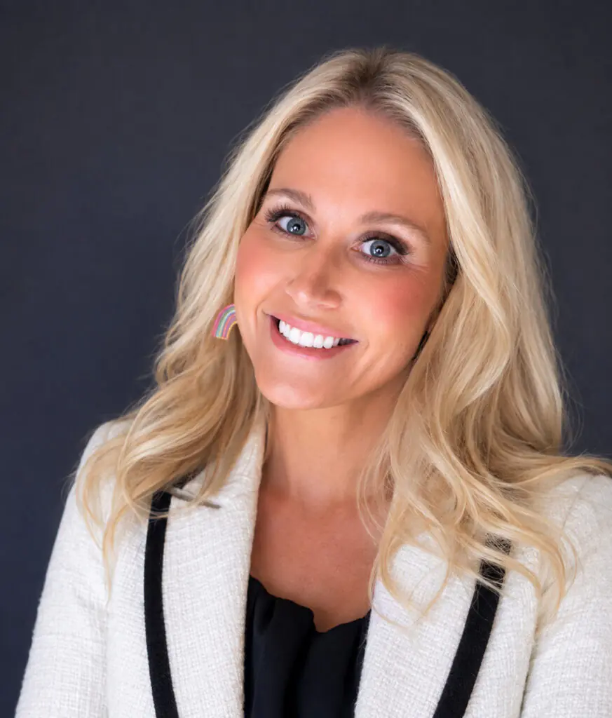 Blonde woman in white jacket smiles against dark background