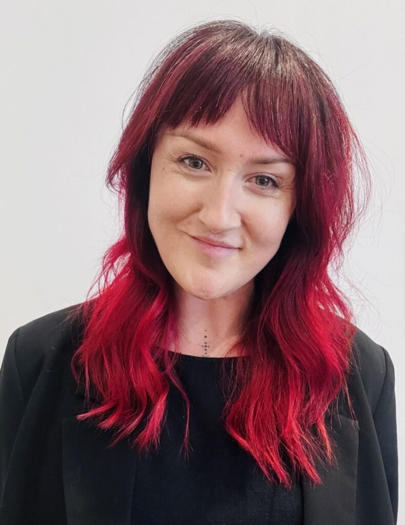 Woman with dark pink hair and fringe smiles against white background
