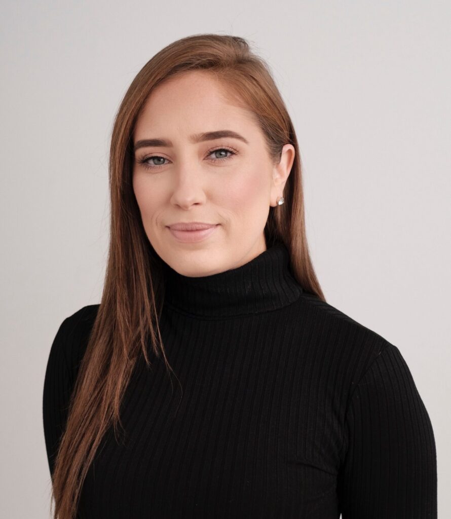 Woman with long brown hair and black turtleneck smiles against a grey background