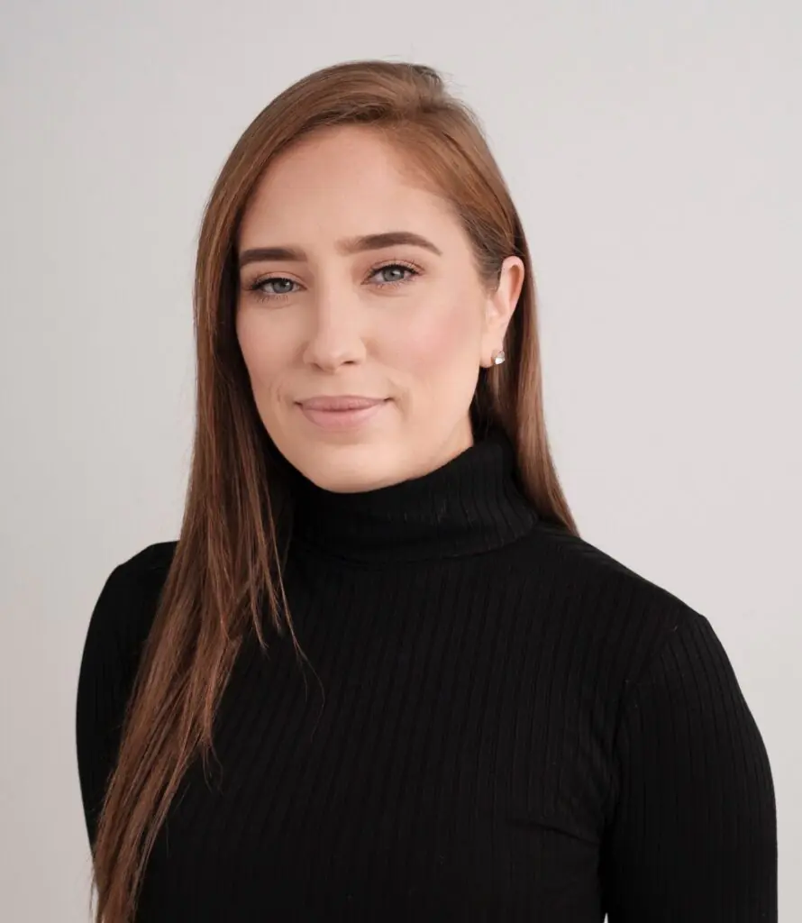 Woman with long brown hair and black turtleneck smiles against a grey background