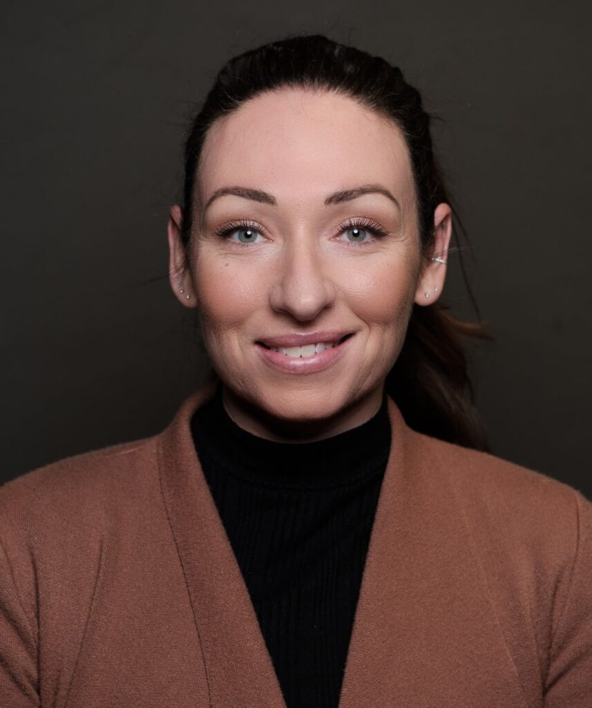 Woman with dark brown ponytail in brown jacket smiles against a dark background