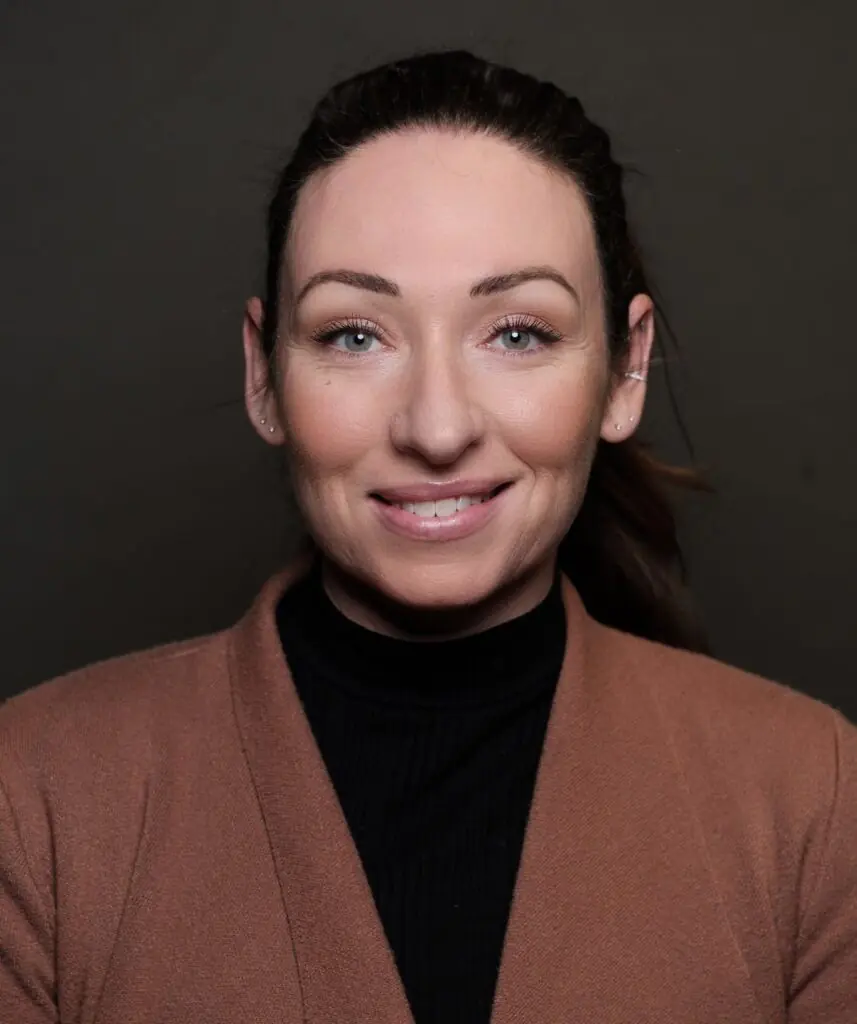Woman with dark brown ponytail in brown jacket smiles against a dark background