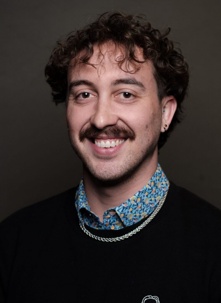 Person with short curly hair and moustache smiles against a dark background