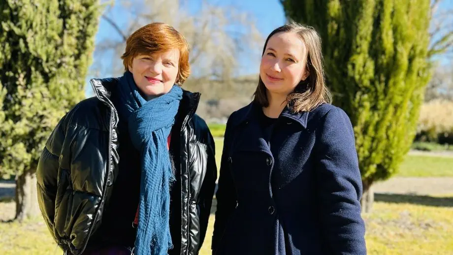 One red-haired and one brown-haired woman in coats smile against a garden background